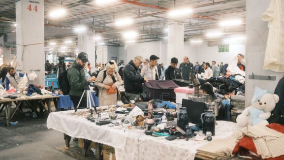 Shoppers browsing tables of secondhand electronics and household items at a busy indoor flea market