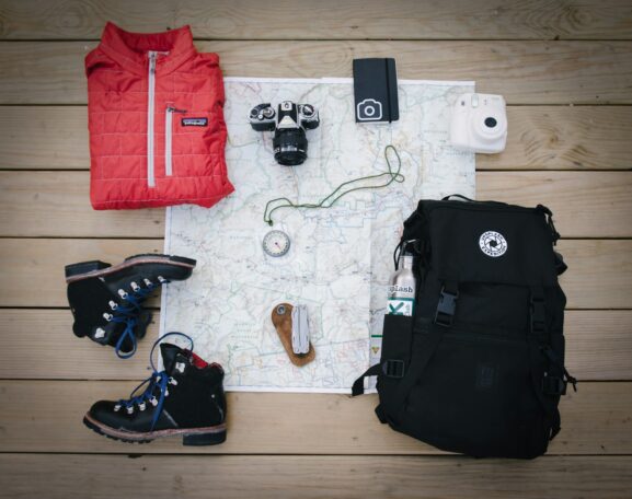 Travel gear laid out on a map: hiking backpack, boots, red jacket, instant camera, compass and watch